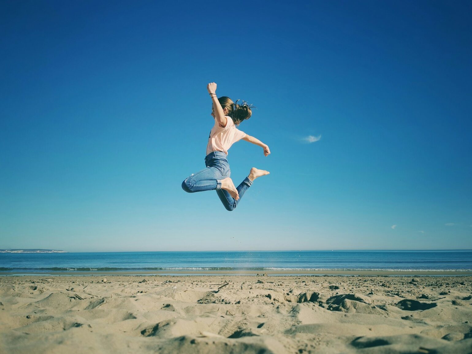 woman in white tank top and blue denim shorts jumping on beach during daytime